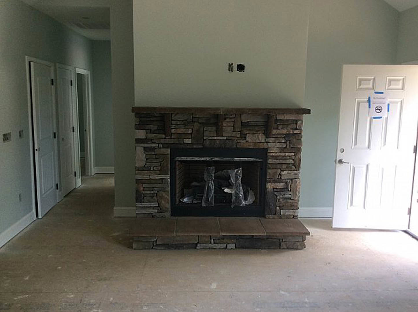 Black fireplace with plastic bags covering the hearth, white door with a sign, brick wall, hardwood floor, and white ceiling in a residential living room.