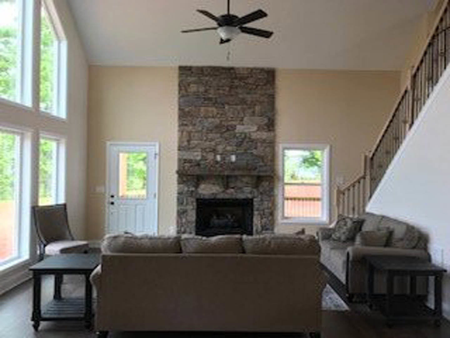 Living room with stone fireplace, ceiling fan with light, upholstered couch with patterned pillows, accent chair, wooden coffee table, hardwood floors, neutral walls