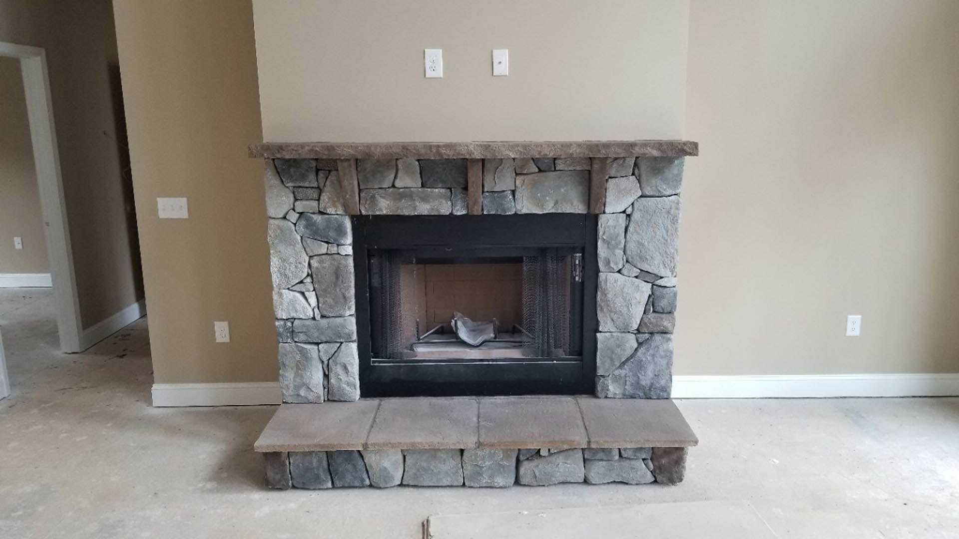 Stone fireplace with black metal frame, built-in hearth shelf, white electrical outlet on adjacent wall, hardwood floor, and fire screen in a modern living room.