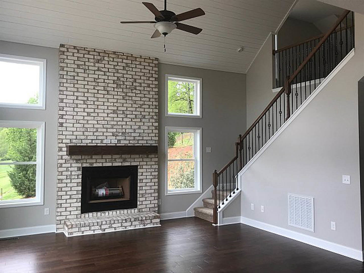 Living room with wood flooring, brick fireplace, white mantel, staircase with wooden railing, ceiling fan with light fixture, neutral walls
