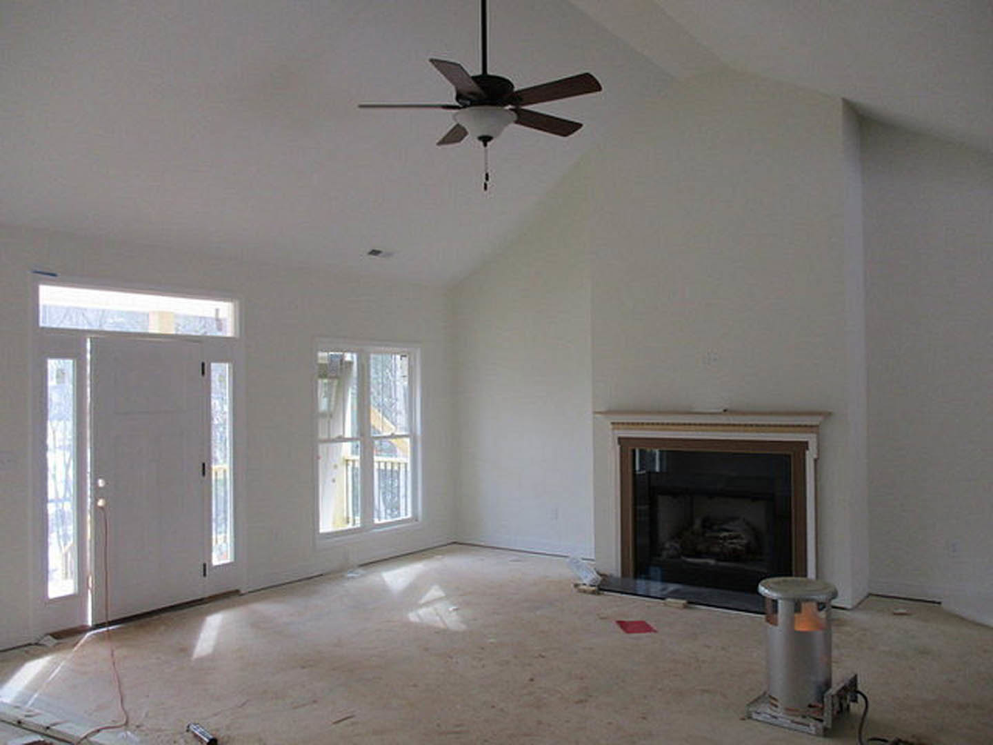 Living room with ceiling fan, glass-enclosed fireplace, white door with glass panes, window featuring metal railing, light-colored plaster walls, and hardwood flooring