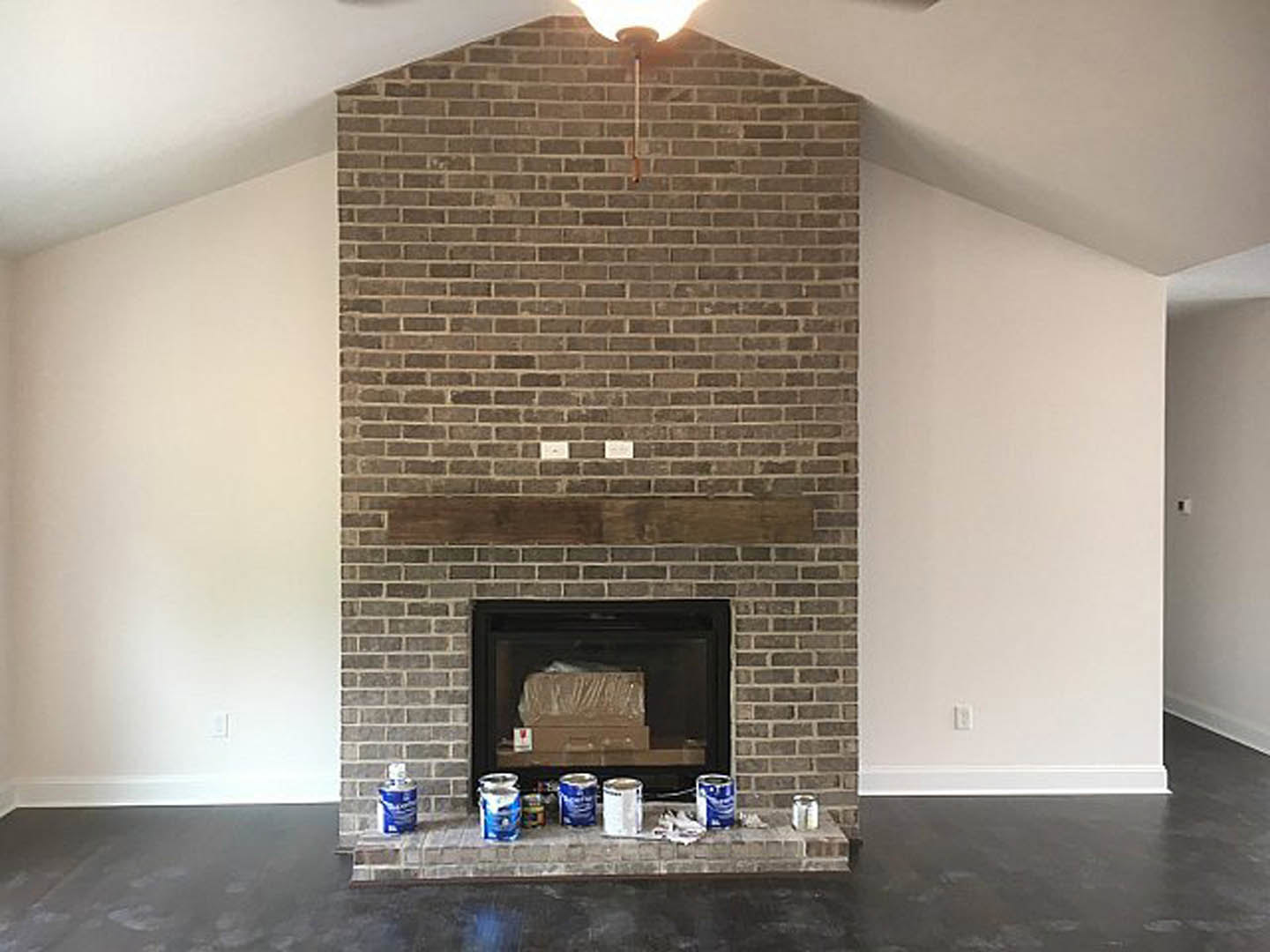 Brick fireplace with wood mantel shelf, paint cans stacked beside hearth, cardboard box placed inside firebox, hardwood floor and neutral painted walls in background.