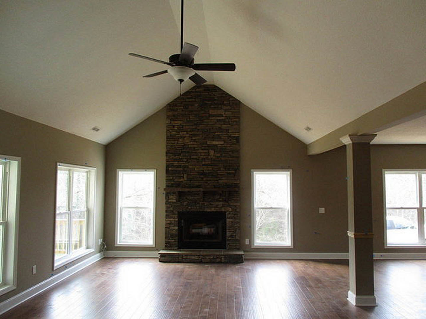 Living room with stone fireplace, ceiling fan with light fixture, wooden flooring, white-framed window, and built-in shelf.