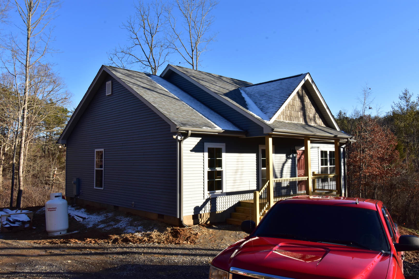 Two-story house with covered porch and white siding, front steps leading to entry, leafless tree in yard, red car parked on driveway, large windows visible