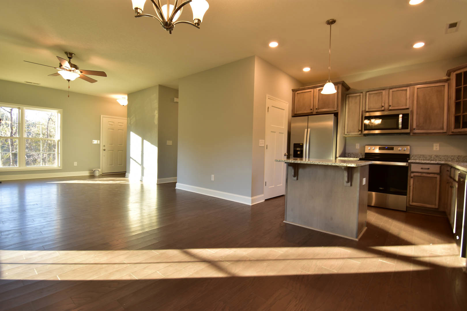 Open-concept kitchen and living room featuring wood cabinetry, stone countertops, stainless steel appliances, ceiling fan, and large windows with views of trees.