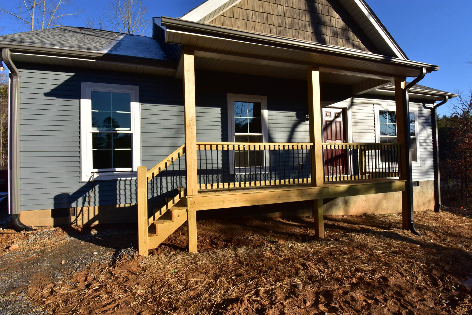 Two-story cottage with light siding, covered front porch, wooden deck and railing, stairs leading to entry, large windows reflecting nearby trees.