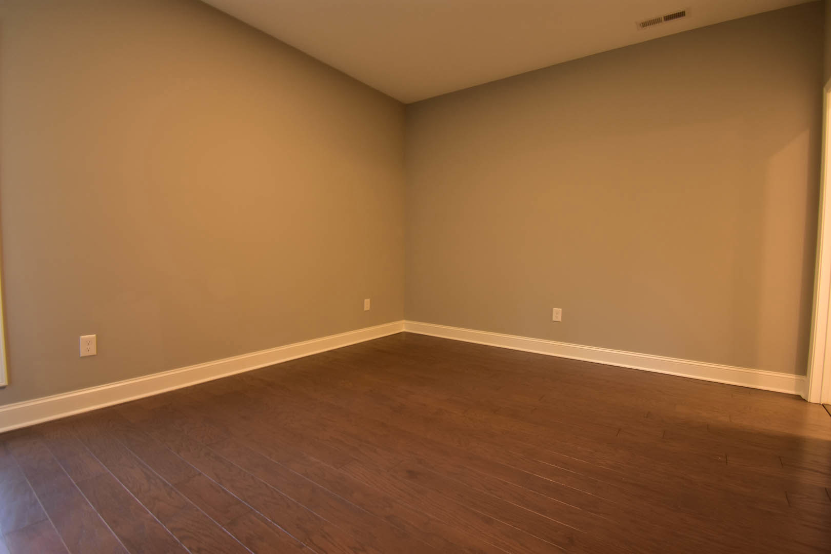 Corner of a room with hardwood flooring, white plaster walls, and a wall vent