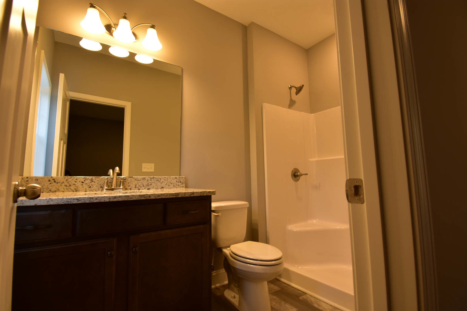 Modern bathroom featuring white porcelain sink and toilet, wall-mounted shower head, light fixture with illuminated bulbs, tiled walls, and close-up view of brushed metal door lock