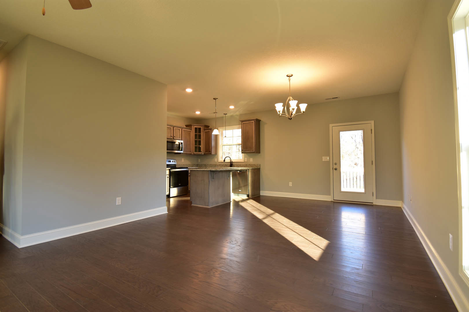 Open kitchen with white cabinetry, hardwood flooring, glass-paneled white door, modern chandelier, sunlight streaming across wooden floor
