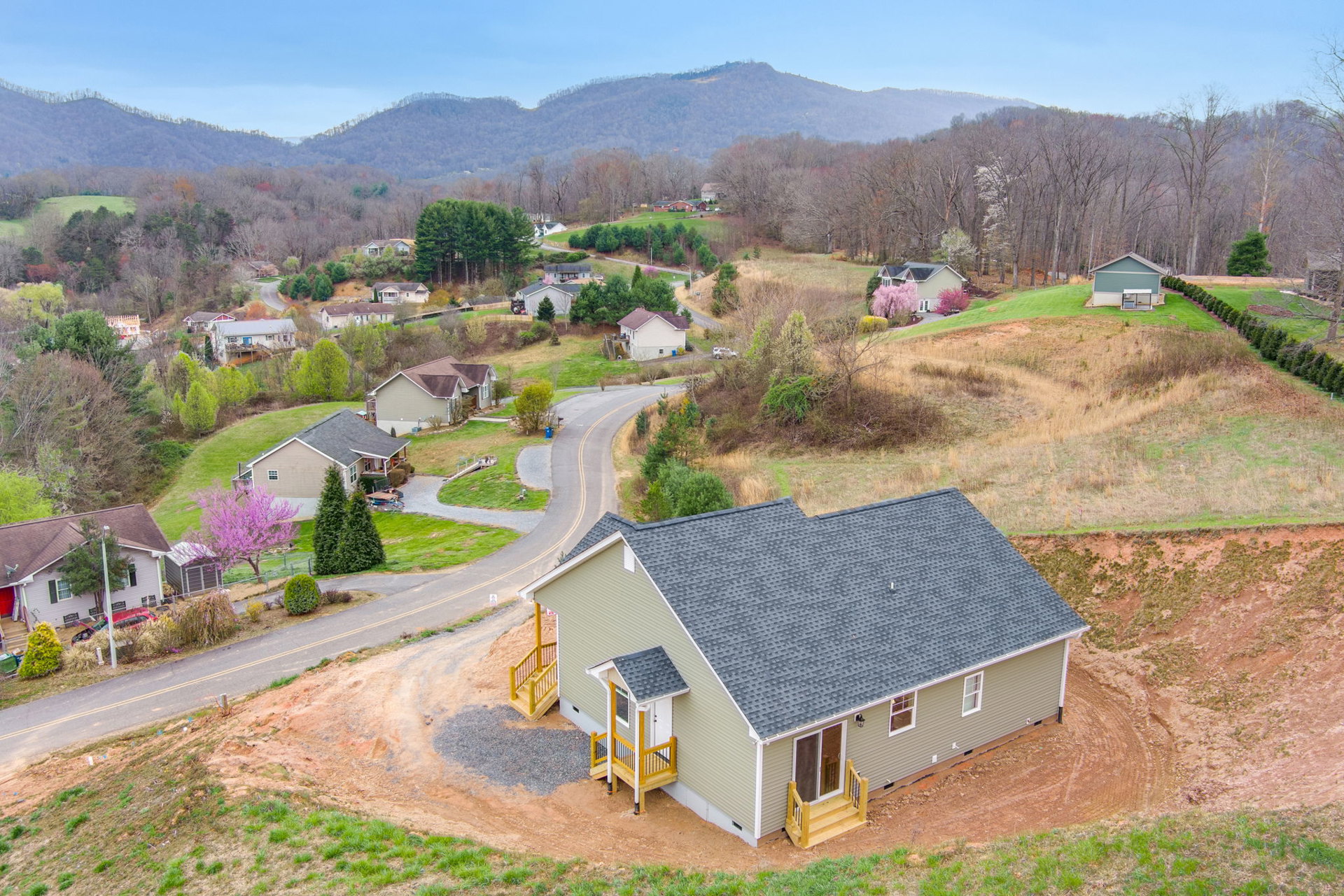 Two-story house with covered porch and front steps, surrounded by green grass, wooden fence, and trees including one with purple flowers and another with pink blossoms, set on a