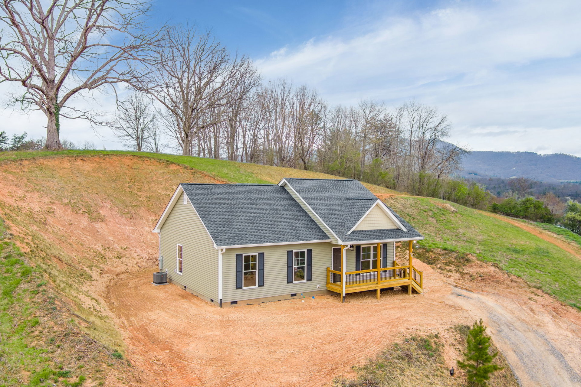 Two-story house with white siding and a porch, yellow railing on the deck, white-framed windows, situated on a dirt hill with grass and a tree nearby under a cloudy sky