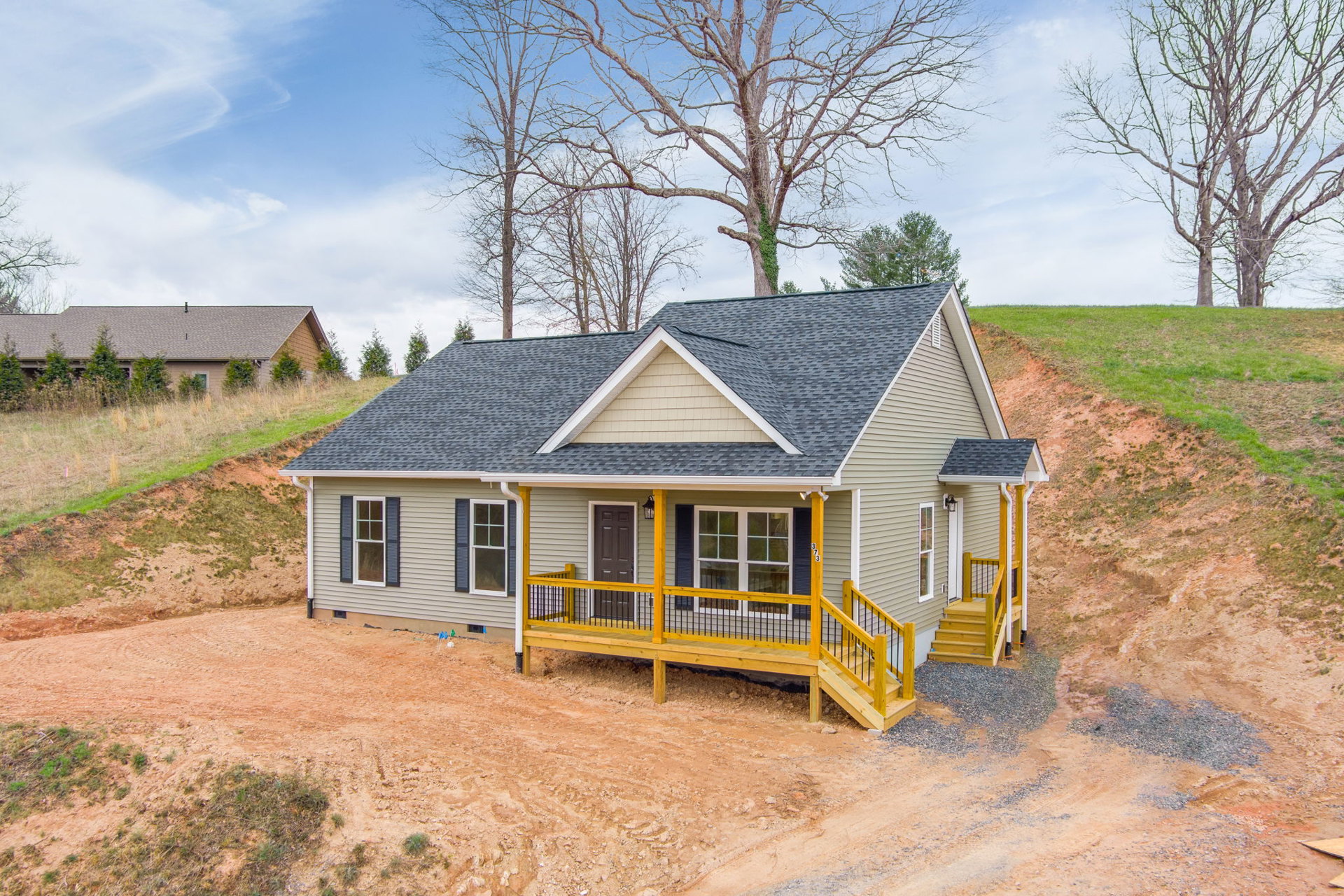Two-story house under construction with exposed wooden deck and yellow railing, surrounded by leafless trees and grassy hill under cloudy sky