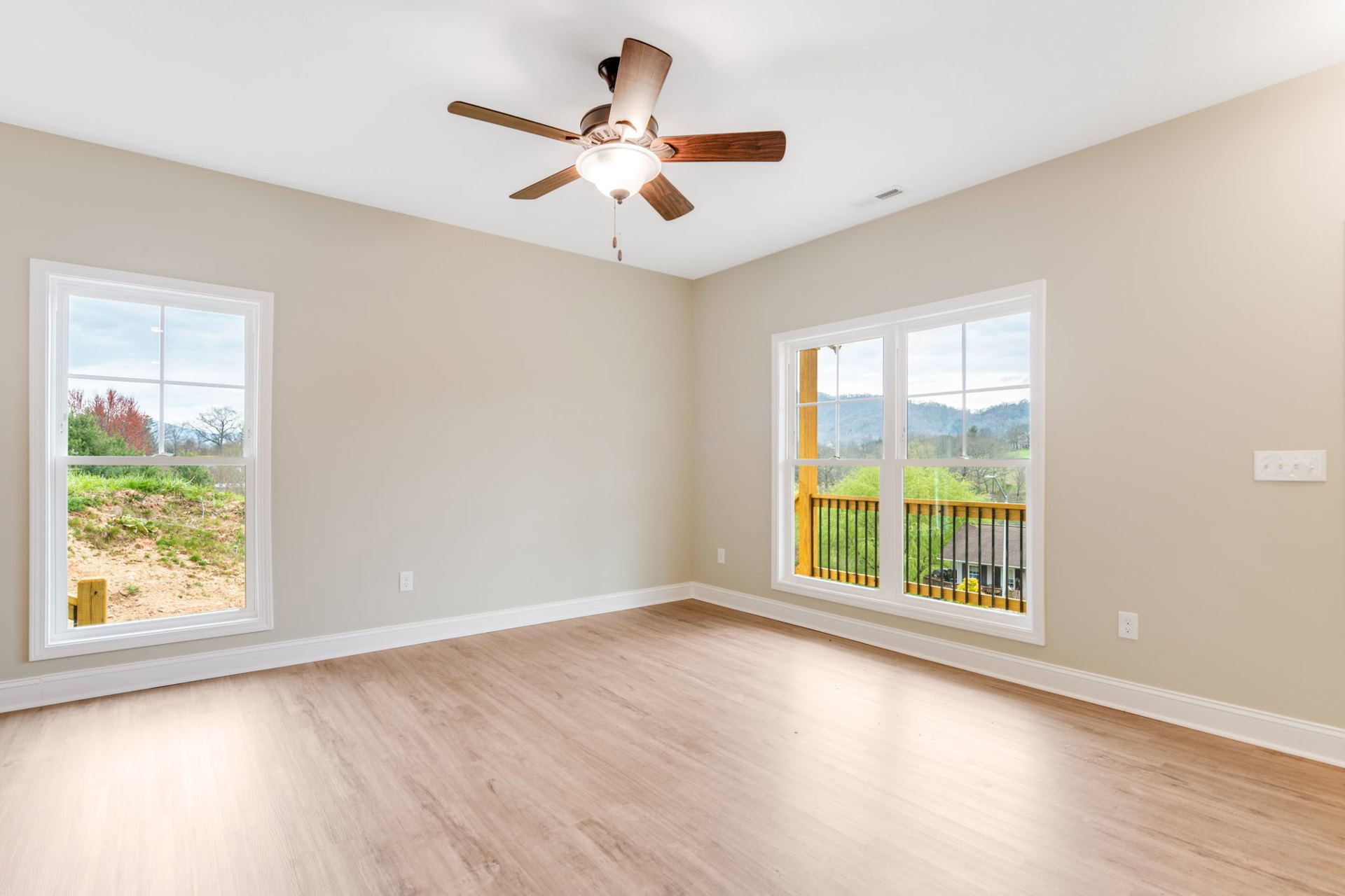 Bedroom with wood flooring, white walls, ceiling fan with light fixture, multiple windows showing views of trees, field, and neighboring house