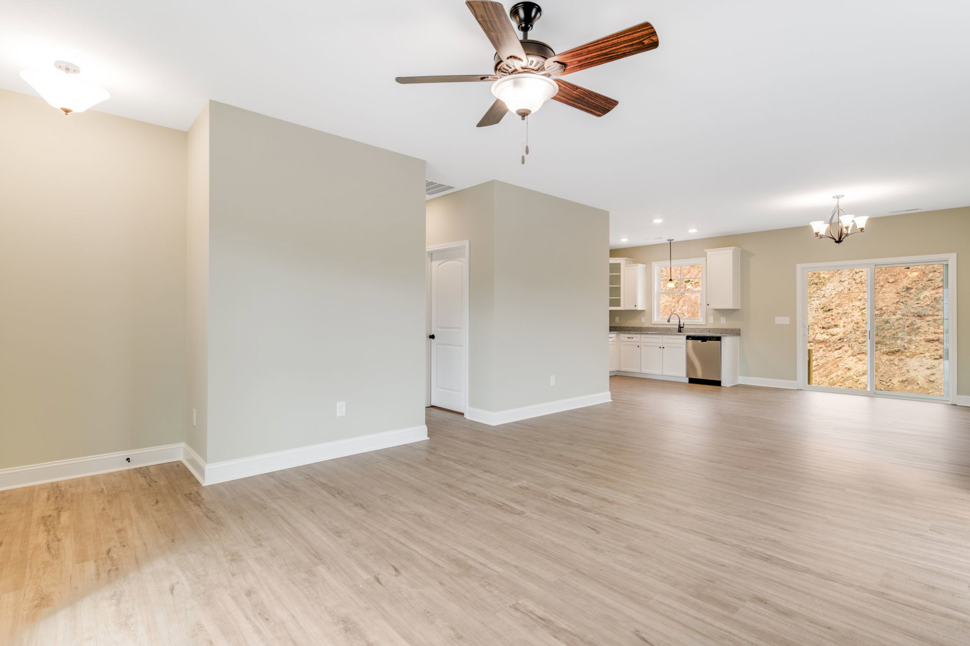 Open kitchen with white walls, wood laminate flooring, ceiling fan with light fixture, window overlooking dirt hill, and black lamp on countertop.