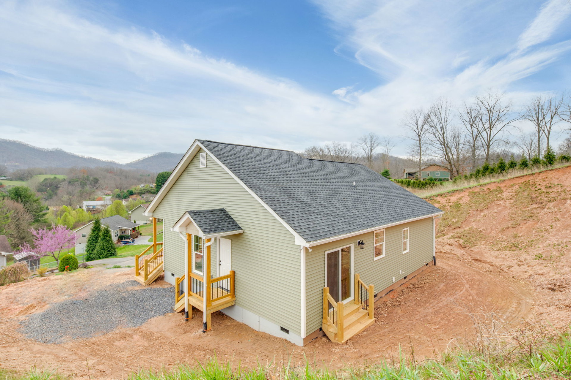 Two-story house with covered porch and wooden stairs featuring metal railings, situated on a grassy hill beside a blooming tree with pink flowers under a blue sky with scattered
