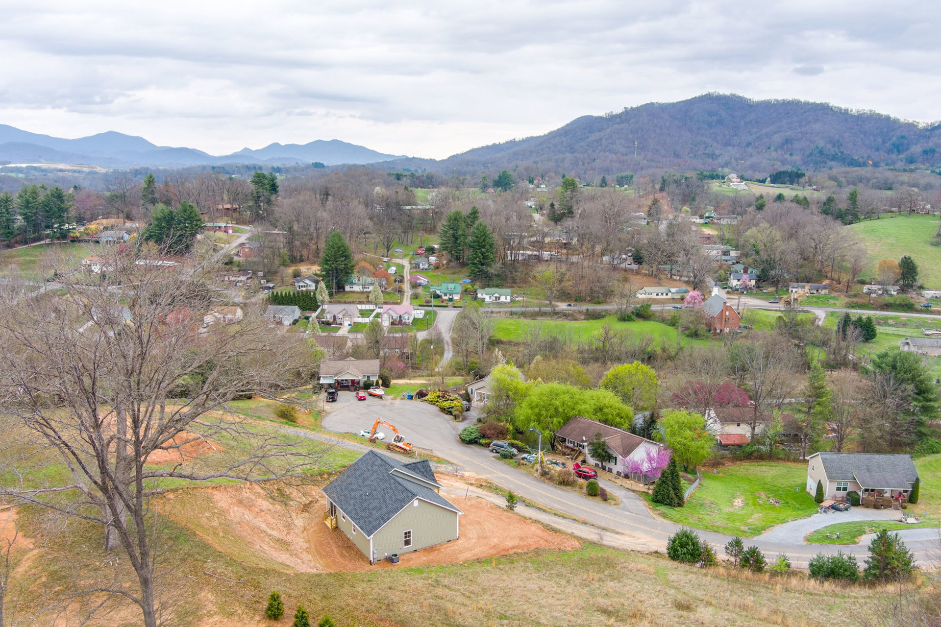 Cluster of houses surrounded by mature trees and grassy landscape, dirt road winding through rural area, cloudy sky above distant hills, open-roof house visible among greenery.