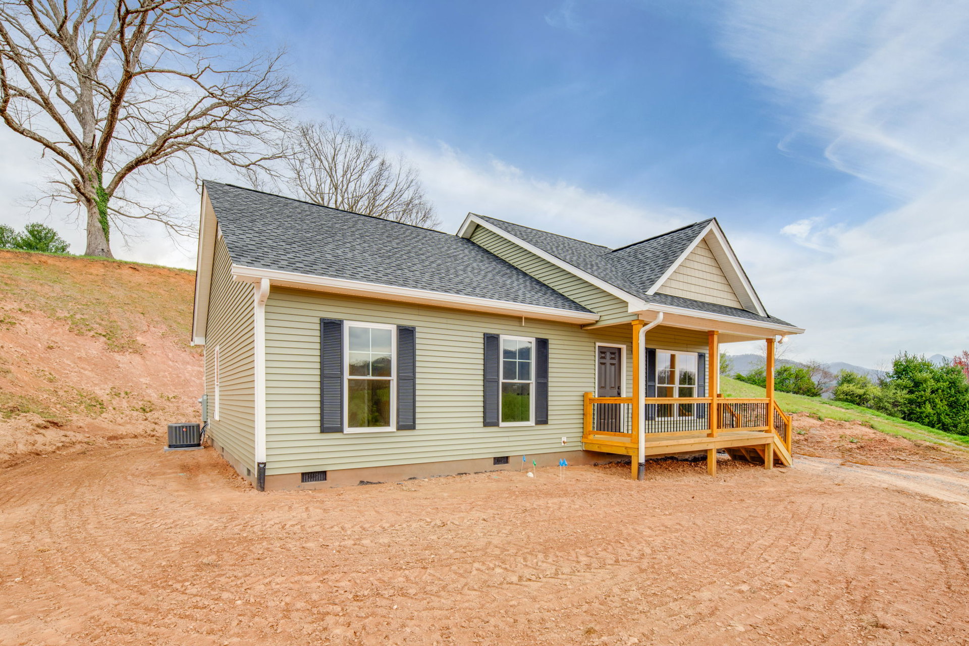 Two-story house with white-framed windows, wooden porch with black railing, dirt hill in front yard, and tree beside exterior under cloudy sky