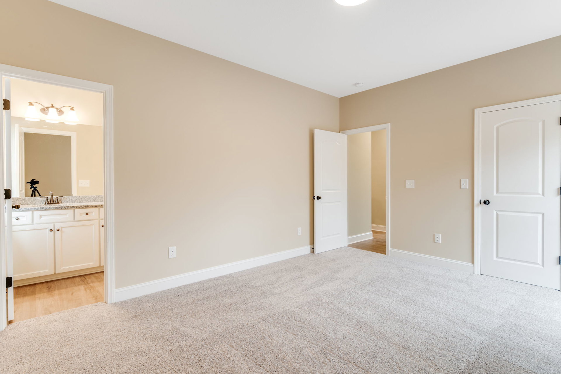 Beige-walled room with open white door featuring black knob, light carpet flooring, and person standing on white surface