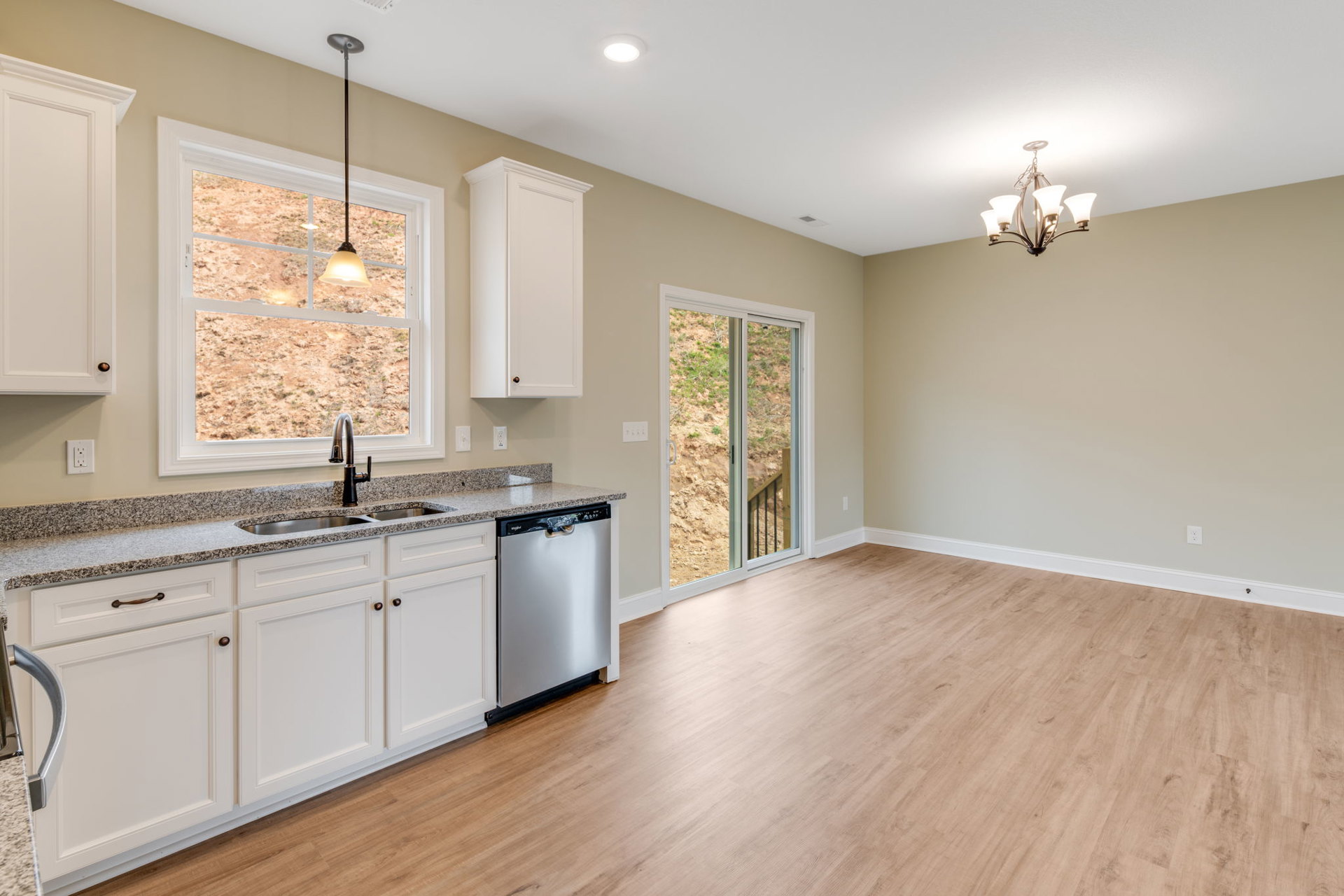 Kitchen with wood flooring, white cabinetry, stainless steel dishwasher, large window above sink, ceiling chandelier, view of staircase through sliding glass door, white door with