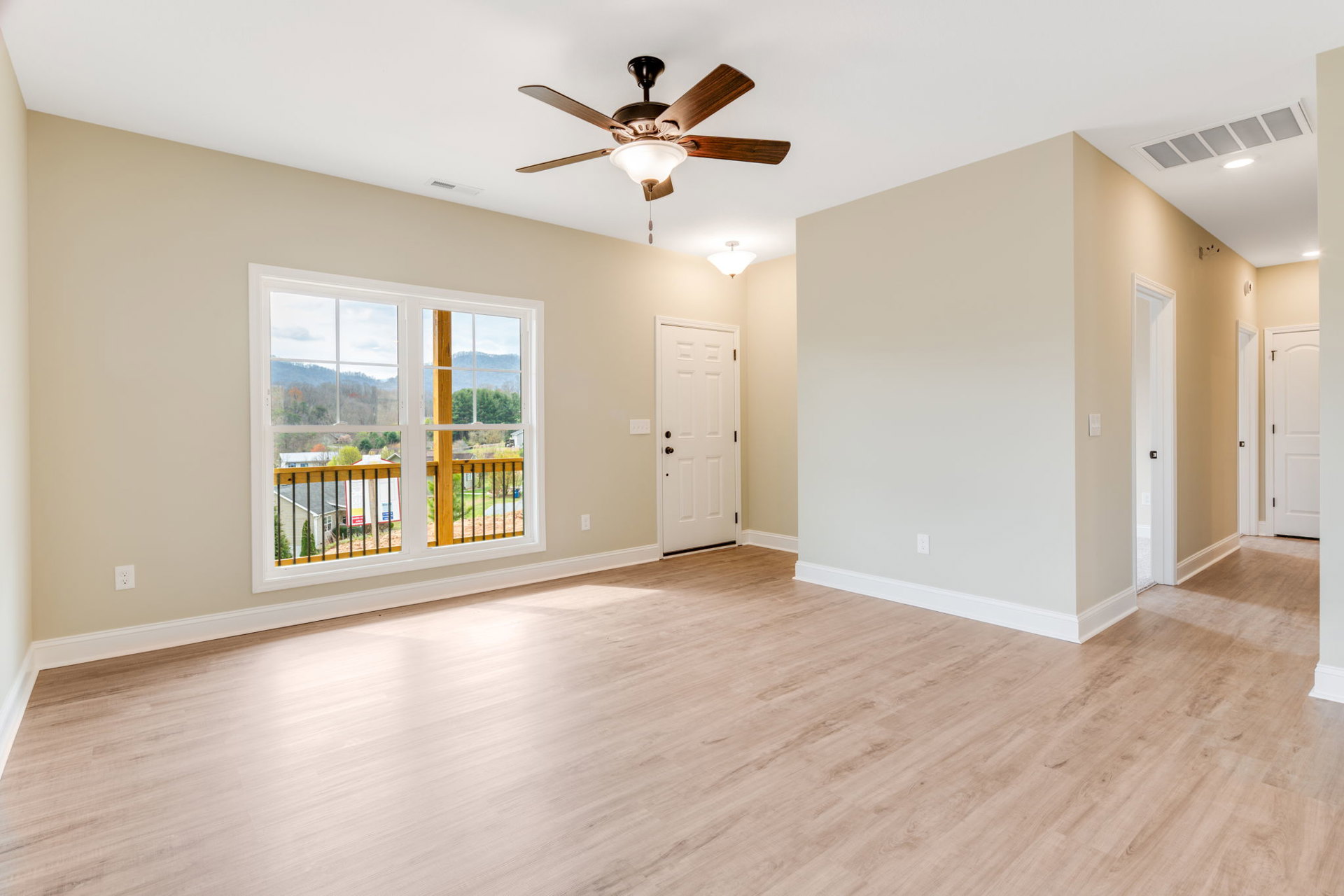 Wood flooring in a bright room featuring a ceiling fan with light, white door with metal doorknob, window overlooking landscape, and close-up of a wood railing