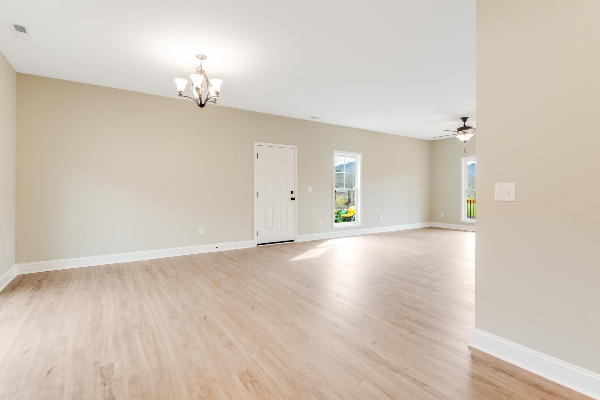 Room with hardwood floor, white paneled door featuring black knobs, white plaster walls, modern chandelier, window overlooking trees and streetlight, and double light switch.