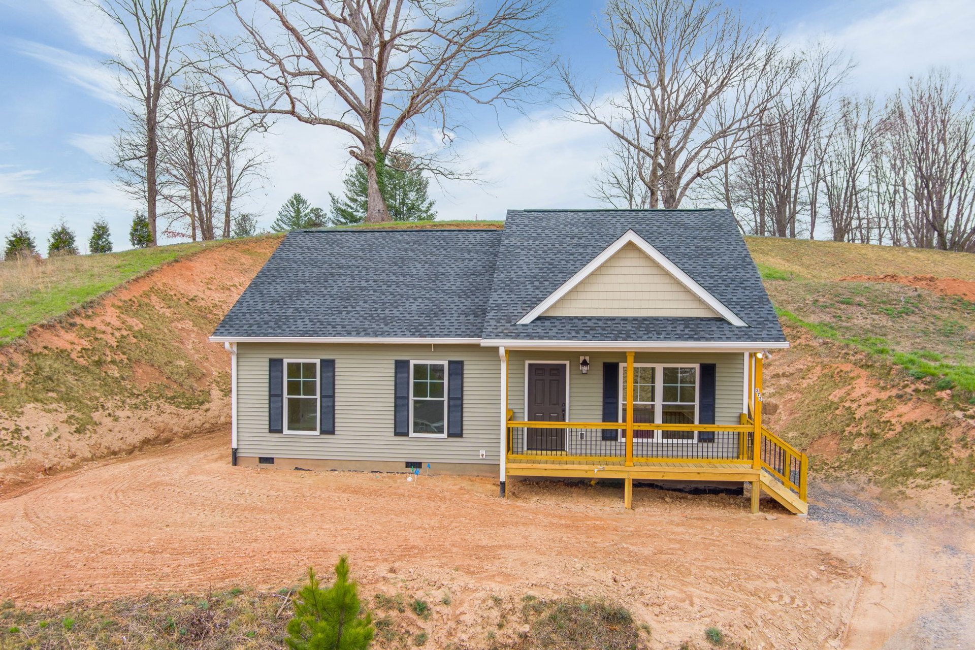 Two-story cottage on a grassy hill with a wooden porch featuring yellow railings, black deck railing, white-framed windows, and a mature tree beside the house under a cloudy sky