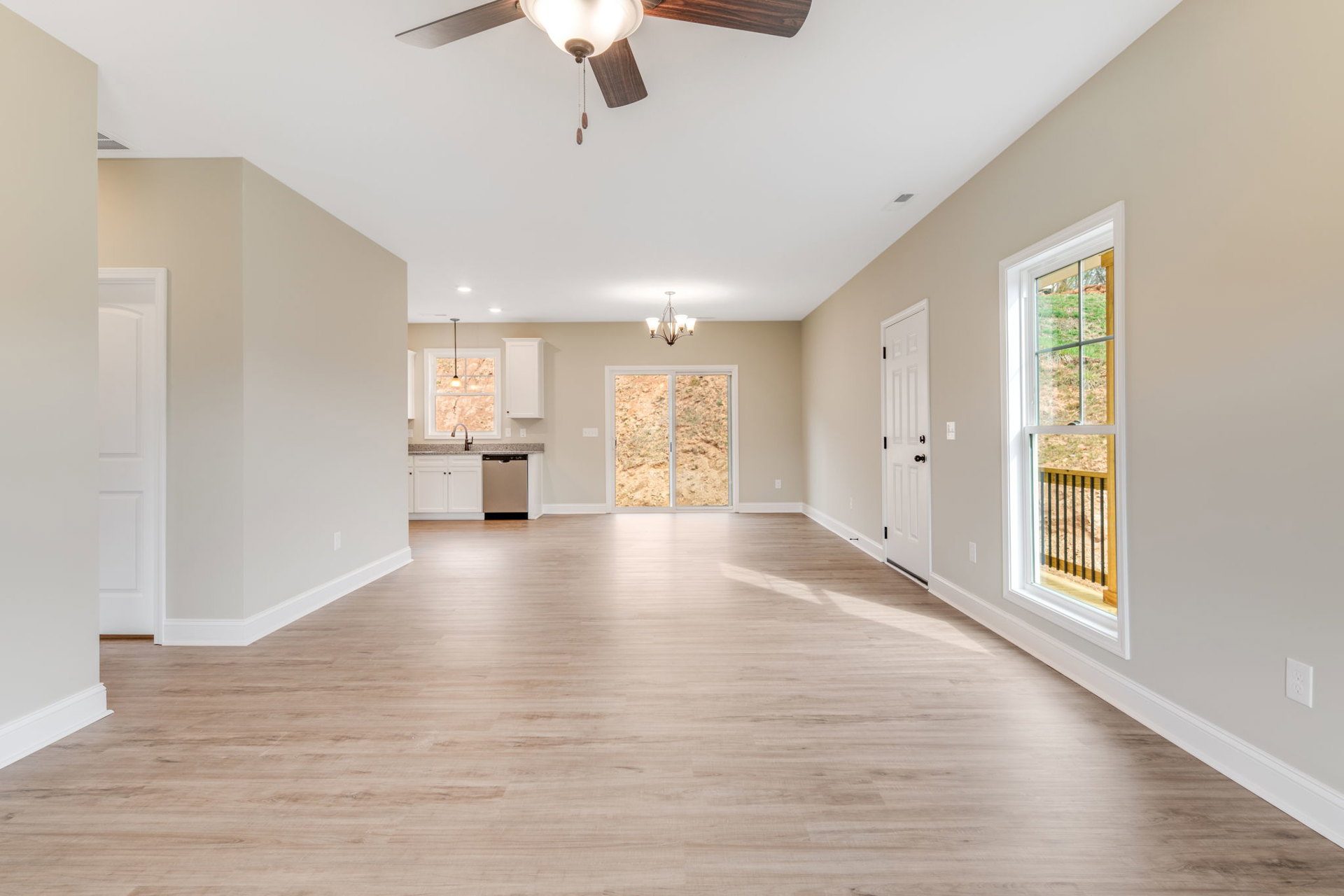 Room with wood laminate flooring, white plaster walls, ceiling fan with light fixture, large window overlooking dirt field, green railing outside, white door with black knobs.