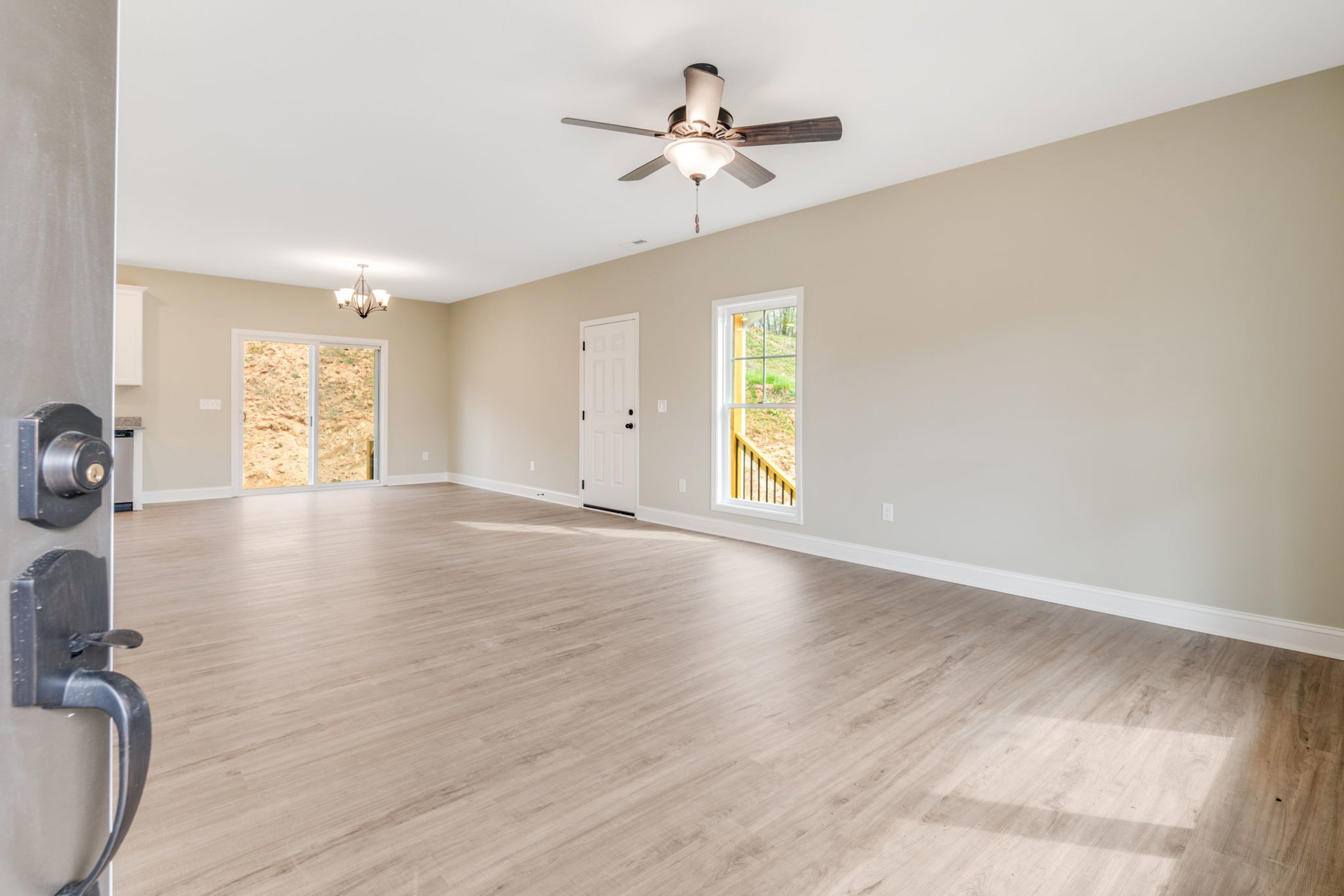 Ceiling fan with light fixture mounted on white plaster ceiling, wood laminate flooring, white door with black knobs, large window overlooking fenced grassy yard and dirt field