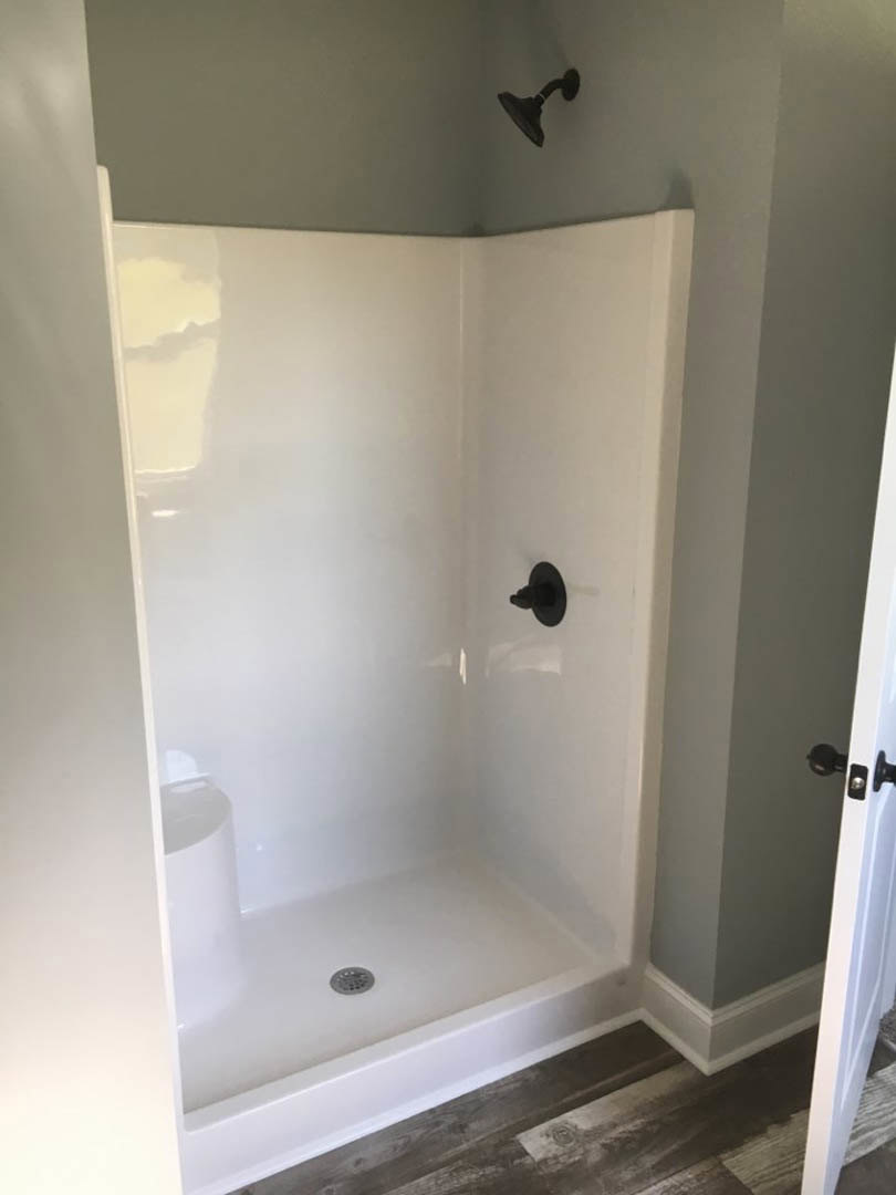 White tile shower with matte black showerhead and handle, wood floor visible in foreground, plaster walls and bathroom door in background