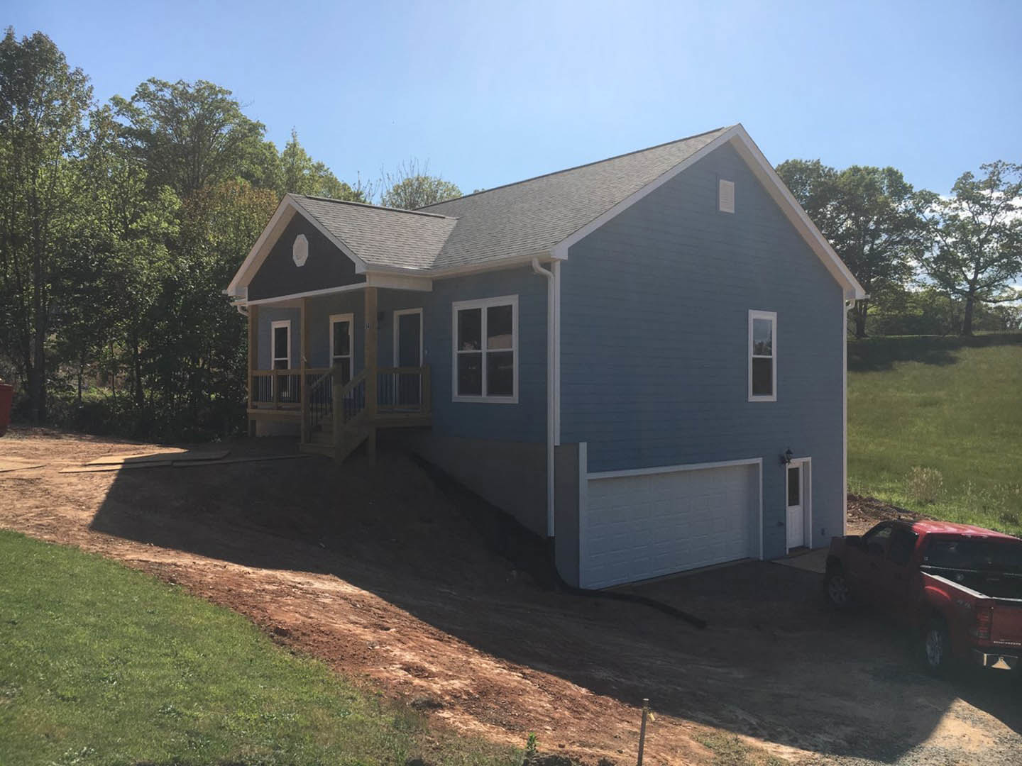 Two-story house under construction with exposed framing, covered porch, attached garage, dirt path leading to entrance, mature trees surrounding property, red truck parked in dirt