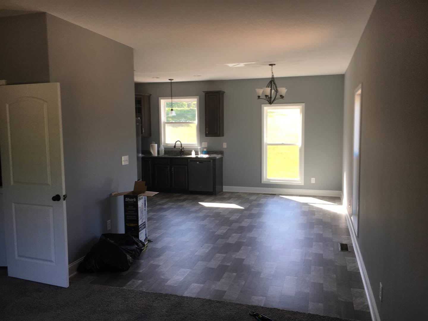 Open room with dark wood floor in rectangular pattern, white walls, white door with black knob, black bag near window, modern chandelier hanging from ceiling