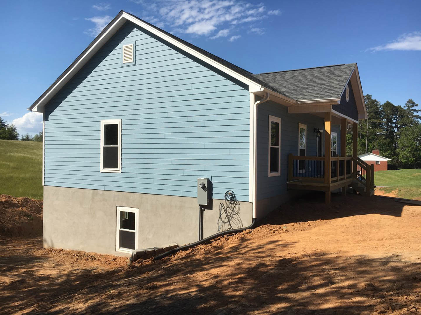 Two-story house with light siding, white-framed windows, covered front porch, elevated wooden deck, black exterior vent, and surrounding trees under a partly cloudy sky