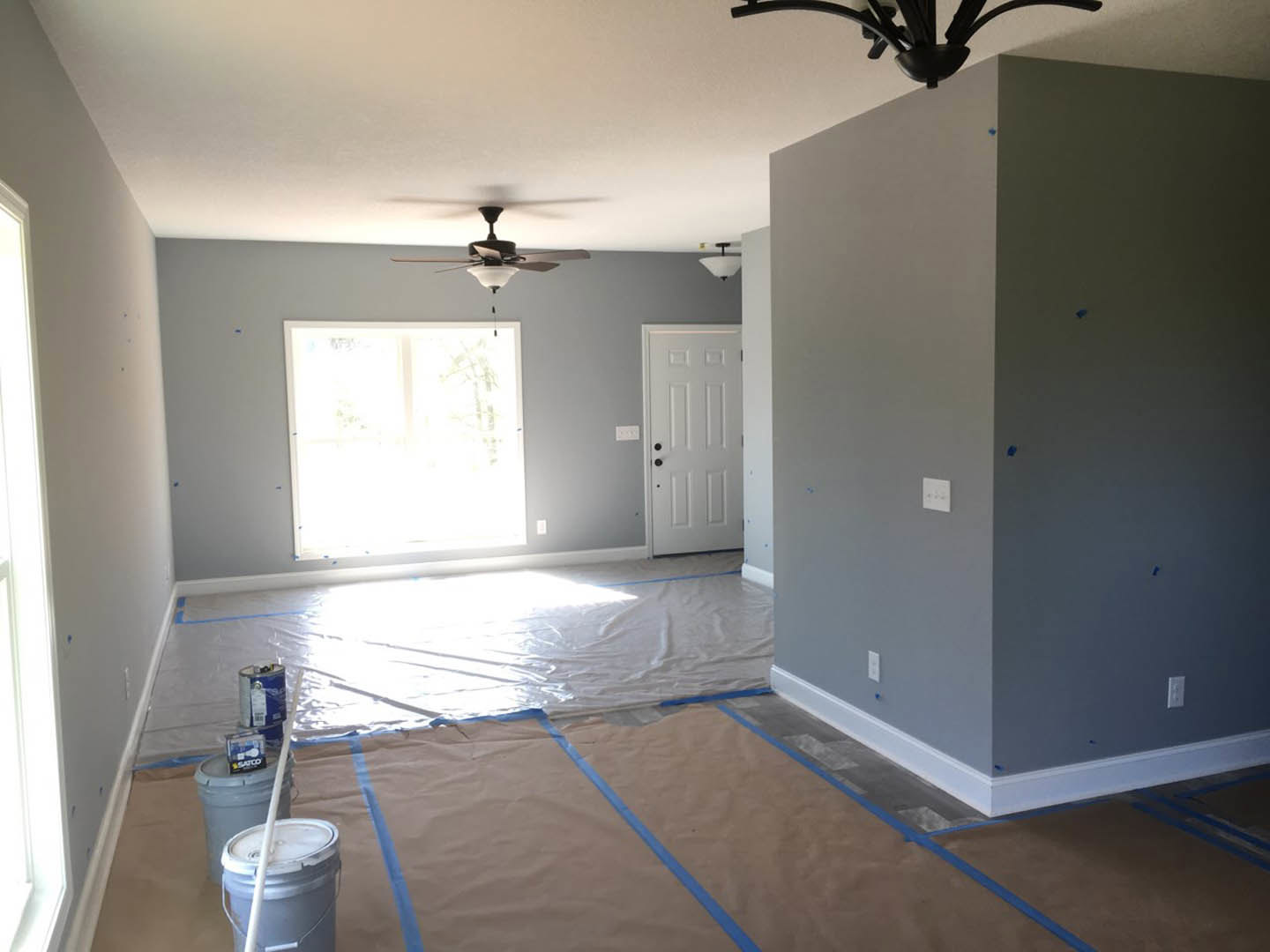 Neutral-toned room with light wood flooring, white walls, ceiling fan, window letting in natural light, white door with black hardware, blue painter’s tape on wall, paint bucket