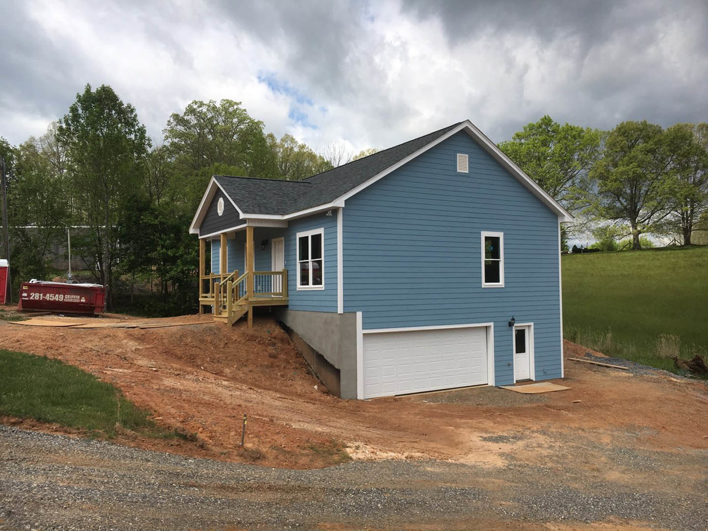 Blue siding house with white framed windows, wooden deck, white garage door under blue roof, driveway, and wooden stairs; red container with white numbers beside home.