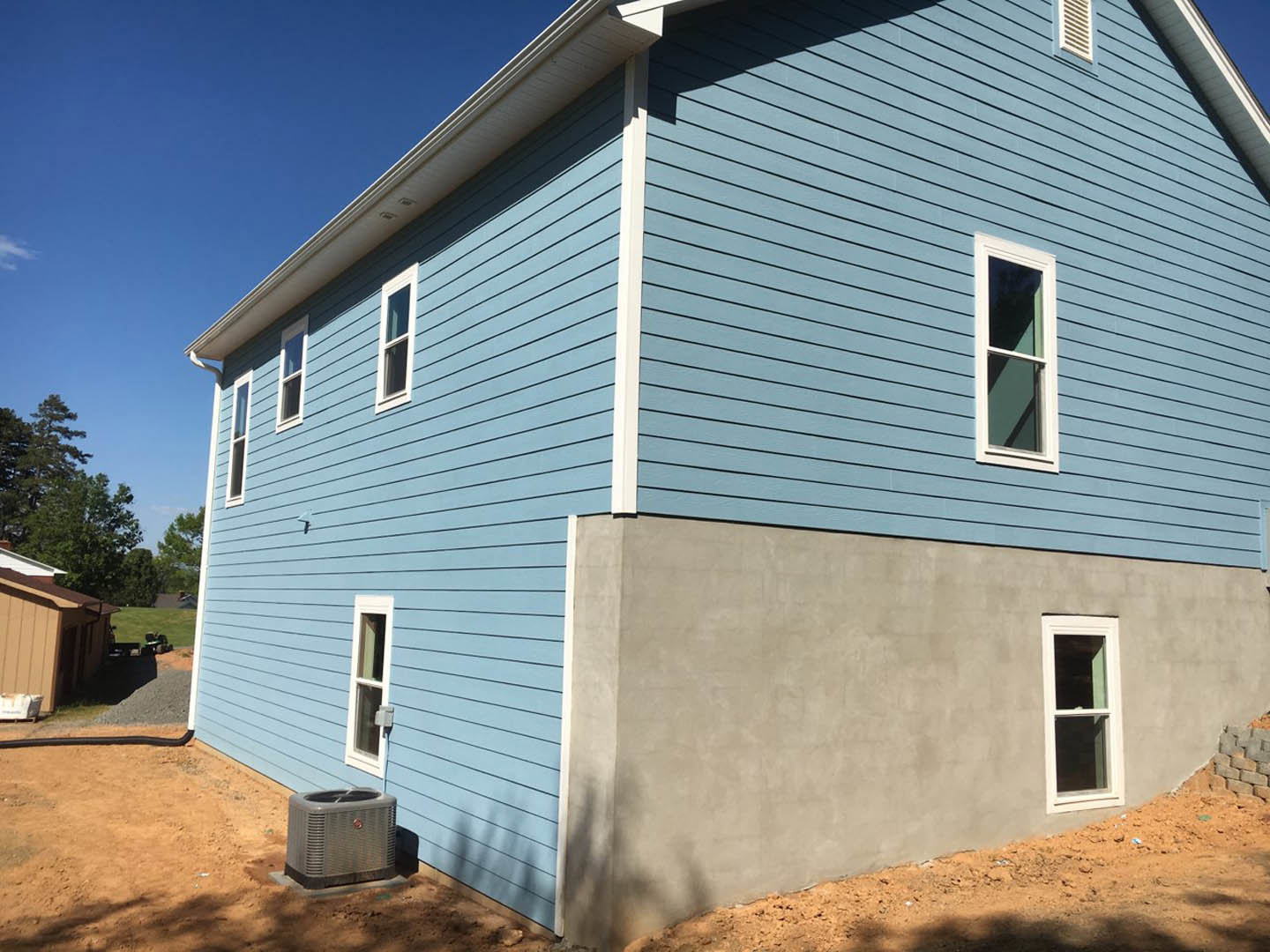 Blue siding exterior with grey foundation, white-framed window, grey air conditioner on concrete, gable roof, clear sky
