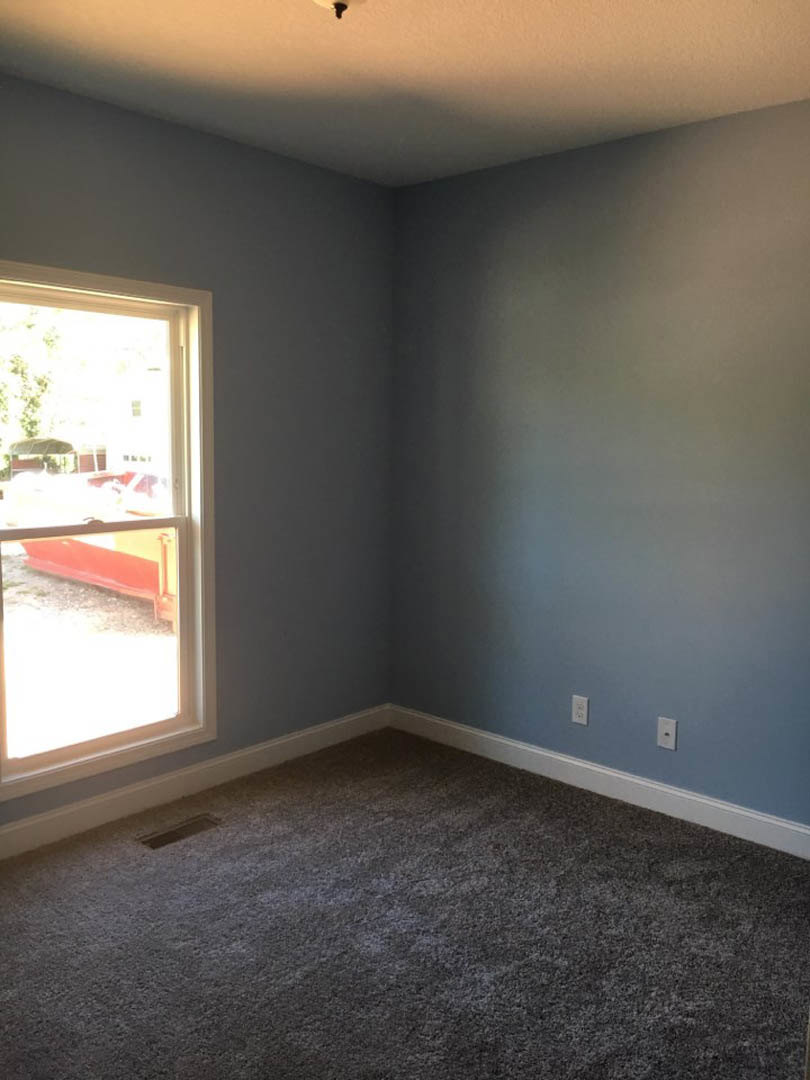 Carpeted room with white plaster walls, ceiling light fixture, and a window framed in red and yellow overlooking a red container outside