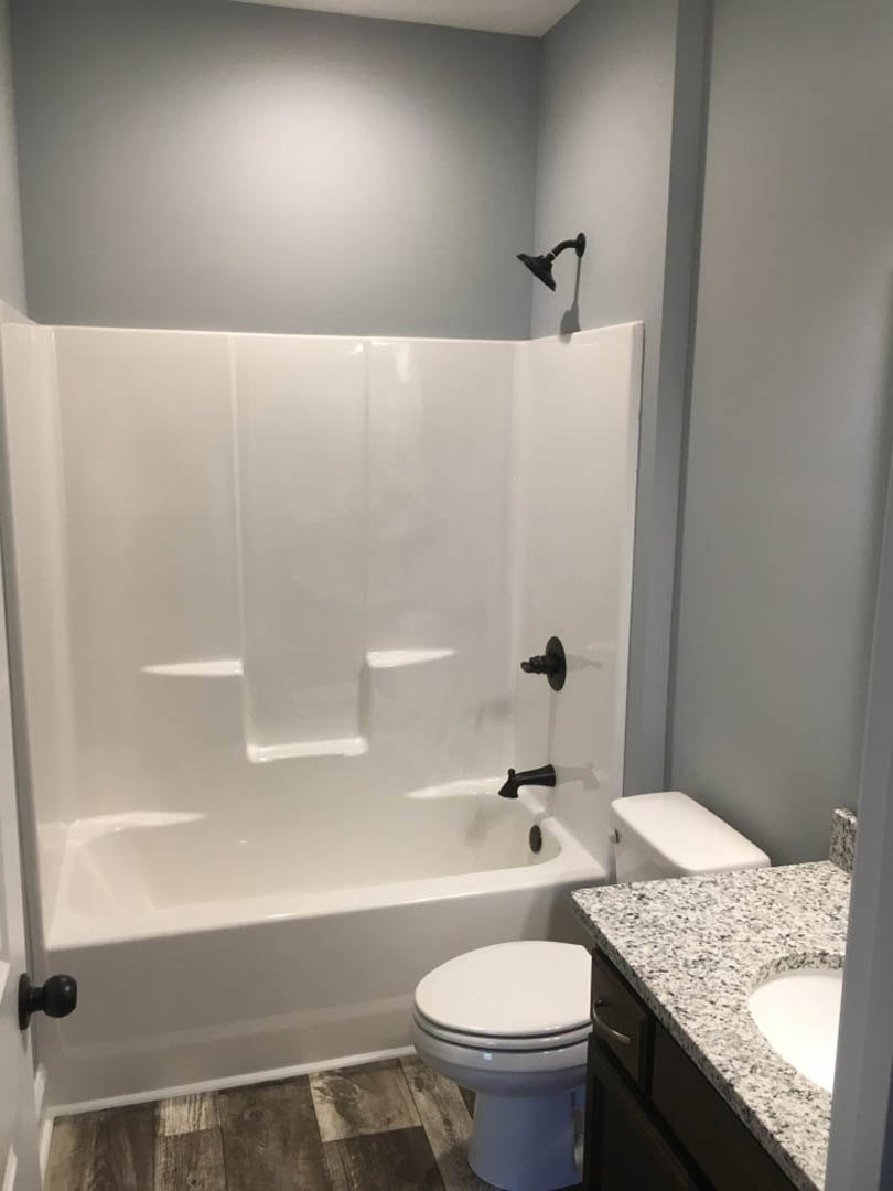 Bathroom with white freestanding bathtub and matching sink, black faucets and shower head, closed toilet seat, light tile walls, black metal towel holder, close-up of silver door