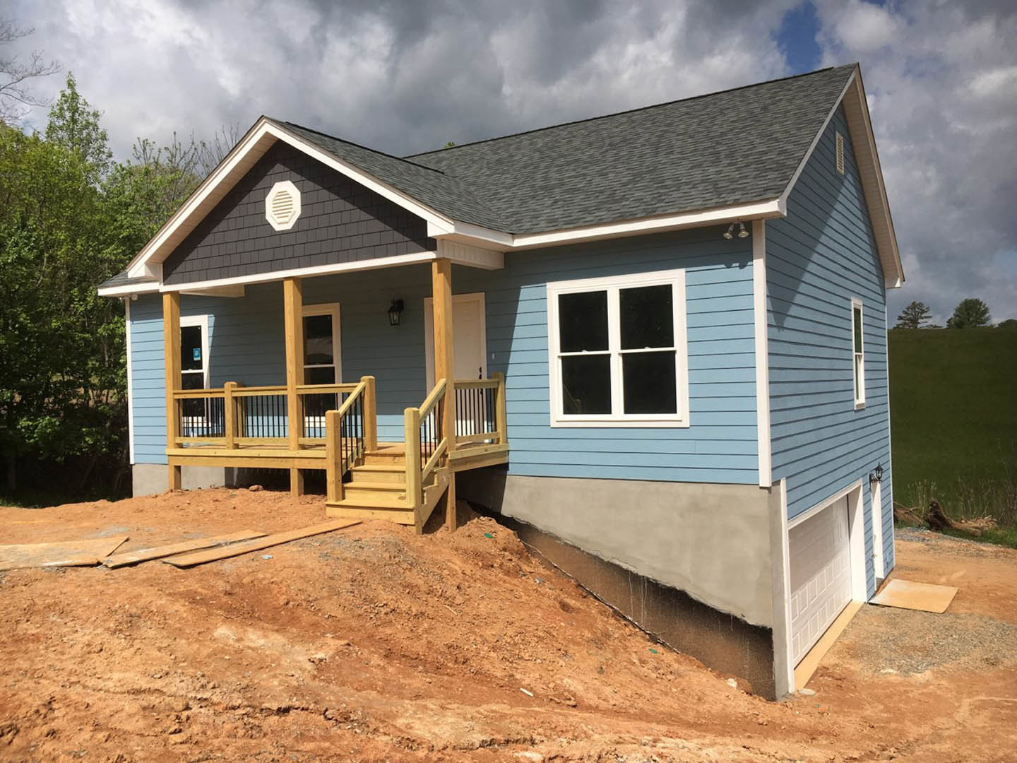 Partially built house with exposed wooden porch, black railing, white vent, large windows, dirt hill in foreground, and surrounding trees under cloudy sky