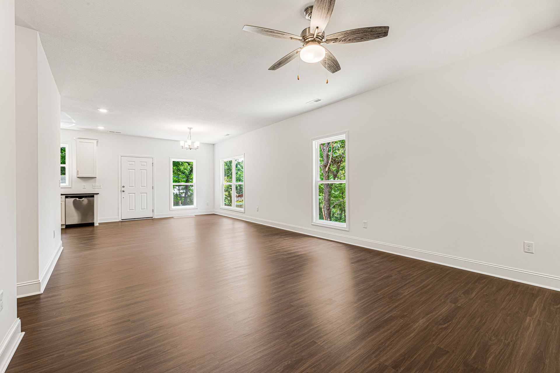 Ceiling fan with light fixture above hardwood floor, white walls, windows showing tree trunks and greenery, white door with silver handle