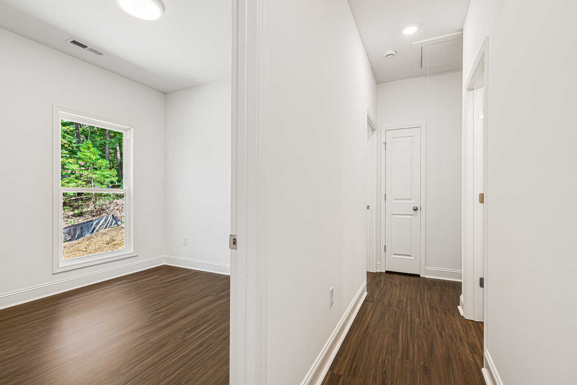 Hallway with white walls, hardwood floor, white door with silver doorknob, window overlooking trees