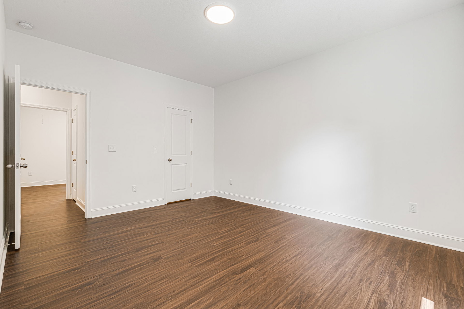 Wood flooring in a room with white walls, a white door featuring a gold doorknob, and a ceiling light fixture