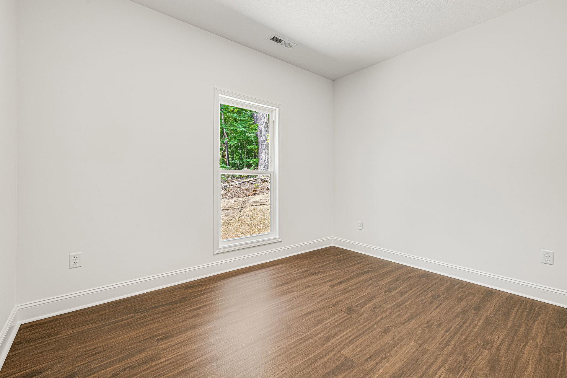 Sunlit room featuring wide-plank hardwood flooring, large window with white trim, white plaster walls, and leafy trees visible outside.
