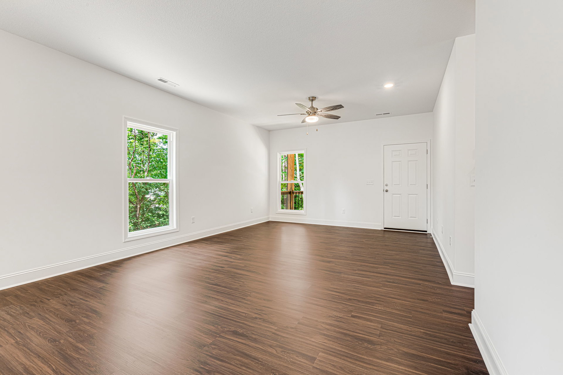 Wood flooring, white walls, ceiling fan with light, large windows showing trees outside, white door with silver knob