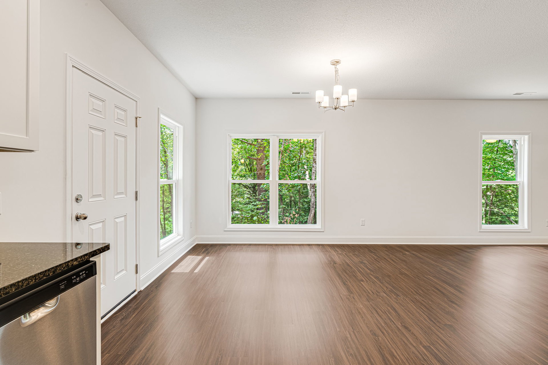 Kitchen with hardwood flooring, white cabinetry, stainless dishwasher with black handle, white door, large window overlooking trees, modern chandelier overhead