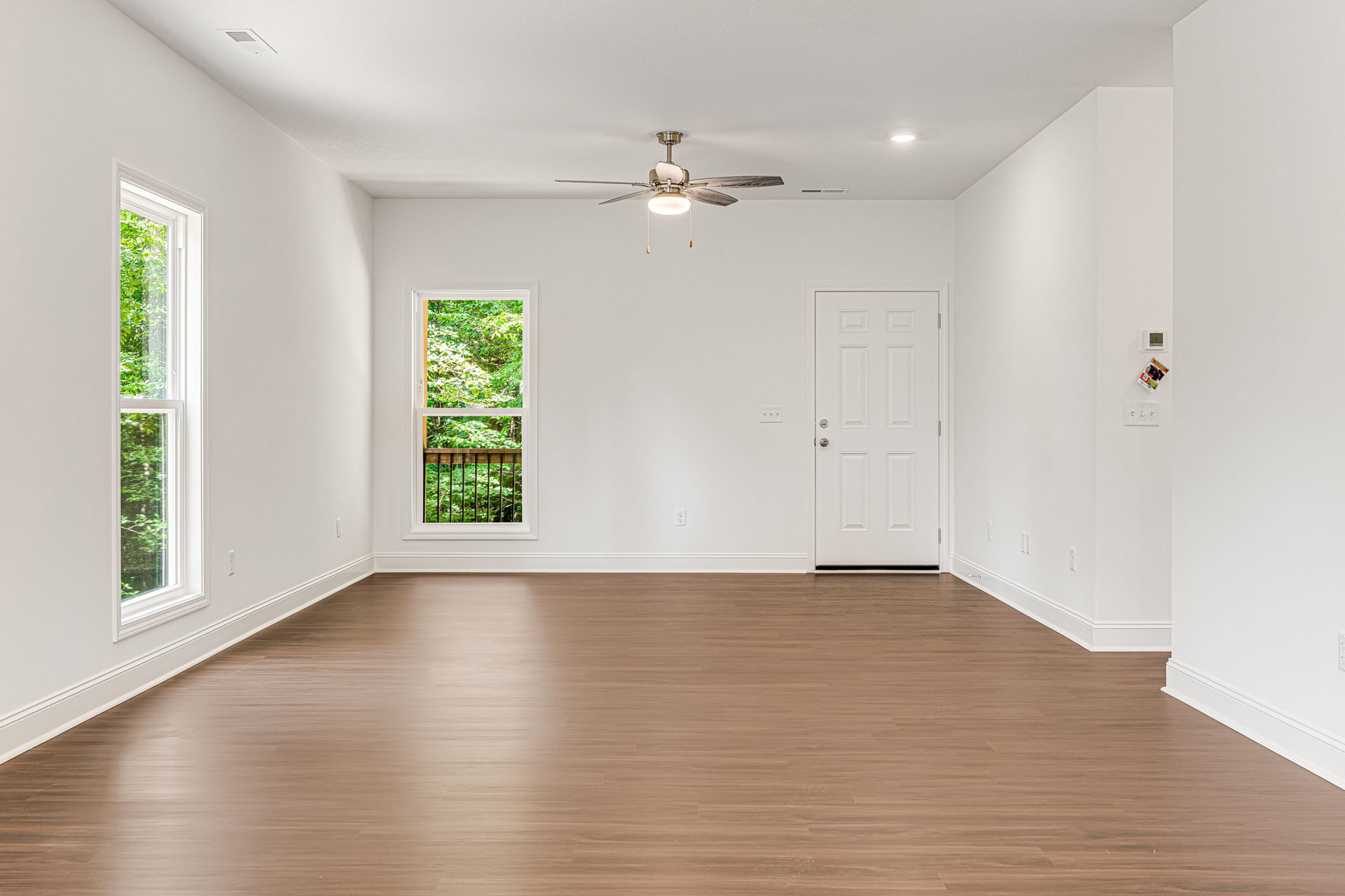 Wood flooring, white door with silver handle, ceiling fan with light, white-framed window overlooking trees and railing