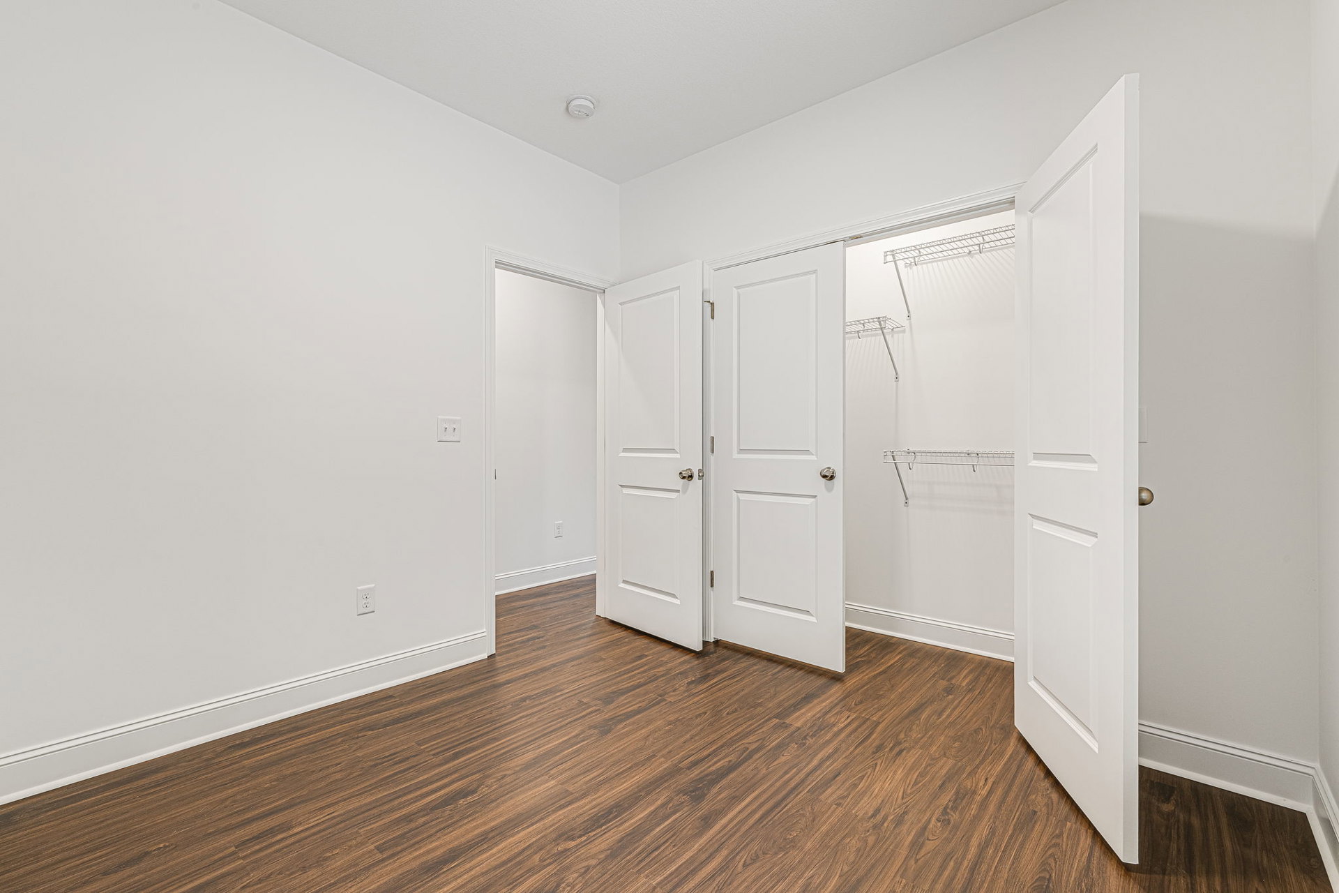 Room with white paneled doors, silver and black handles, light wood flooring, and a white wall-mounted shelf