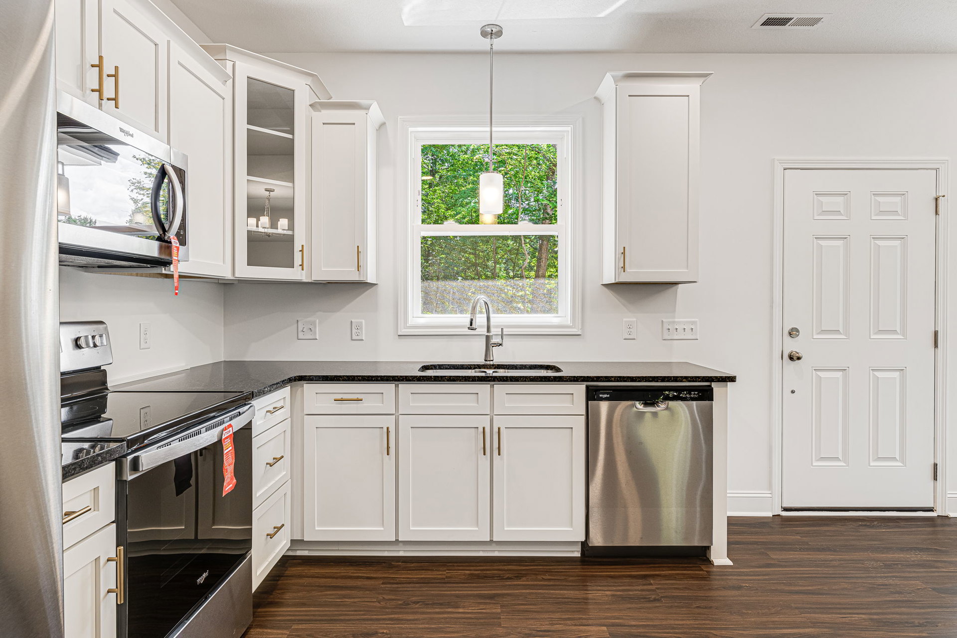 Kitchen with white shaker cabinets, matte black countertops, stainless steel dishwasher, white door with silver handle, window emitting warm light, white door with gold handle