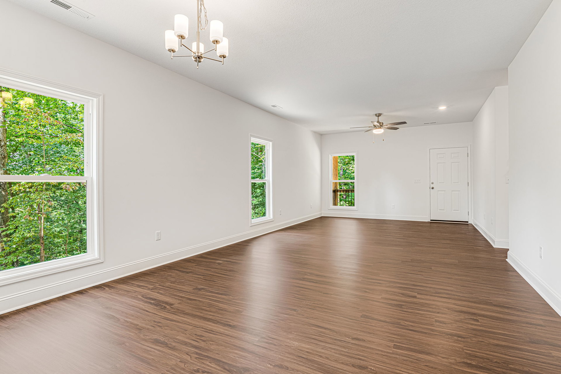 Wood flooring in a bright room with a ceiling fan, white walls, multiple windows showing leafy trees outside, and a white door with a silver doorknob.