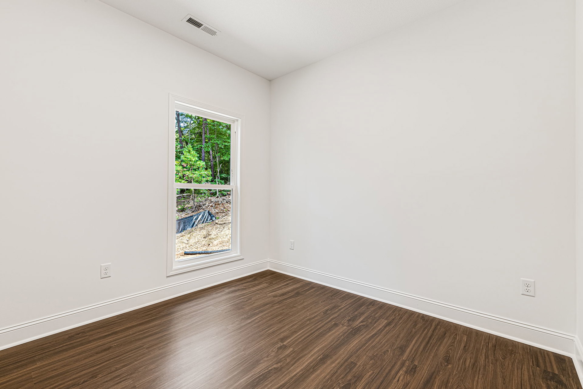 Sunlit room featuring wide-plank hardwood flooring, white plaster walls, large window overlooking forested landscape, floor vent near baseboard, and black tarp partially visible on