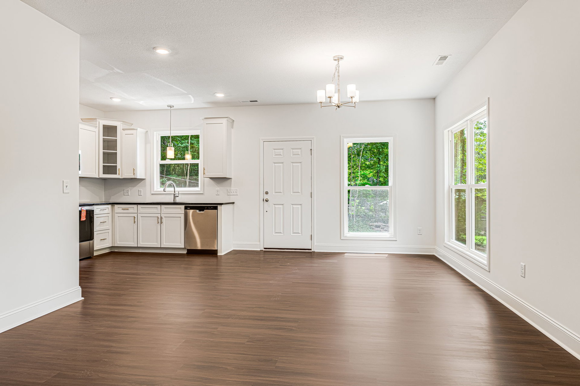White cabinets with gold handles, hardwood floor, black refrigerator, white door, window showing green trees, ceiling light fixture with white shades.
